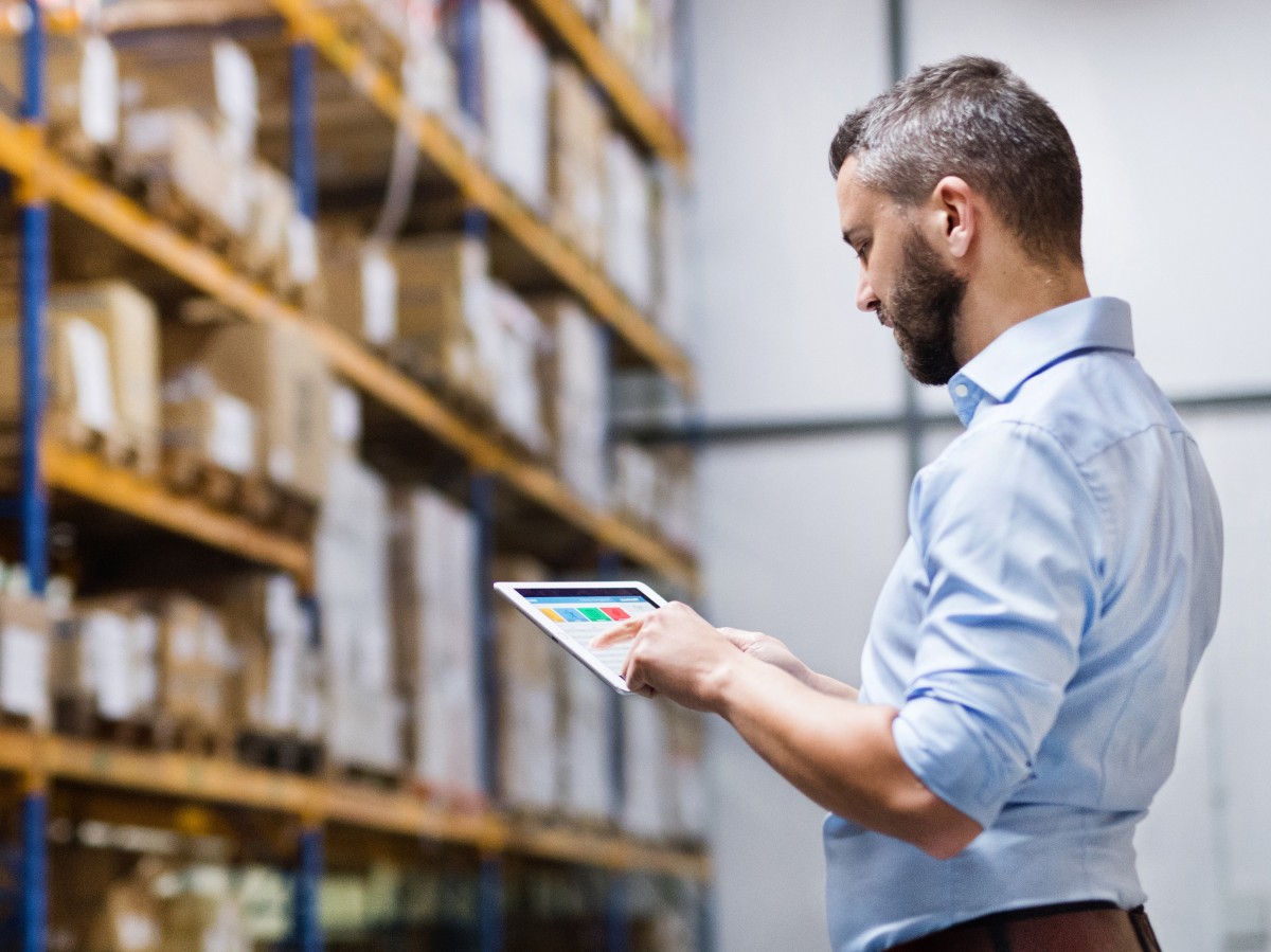Geprüfter Logistikmeister gleicht Projektdaten am Tablet ab mit Warenregal im Hintergrund