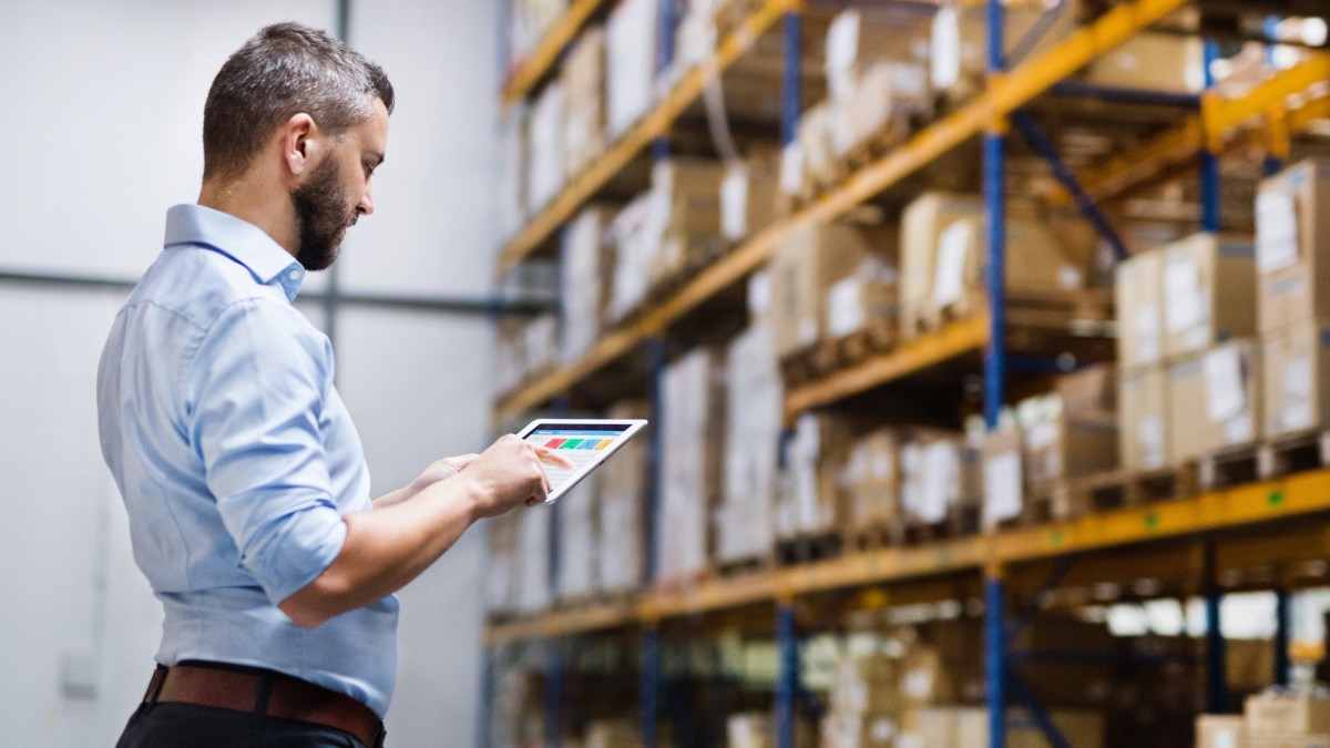 Geprüfter Logistikmeister gleicht Projektdaten am Tablet ab mit Warenregal im Hintergrund